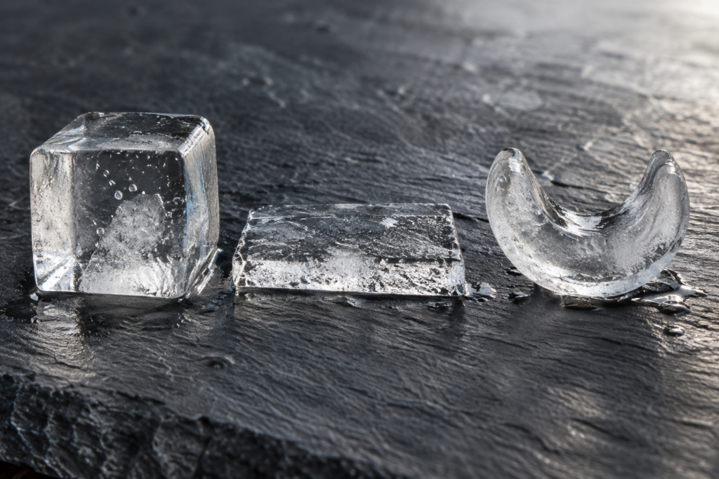 Three pieces of ice lie in a row on dark slate: a full clear cube, a thin flat slab and a hollow crescent.