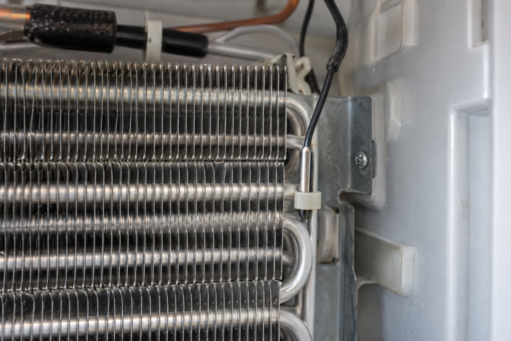 A macro view of a refrigeration cabinet's evaporator block behind a removed interior panel, thin evaporator fins and a small thermistor sensor on a wire clipped beside them.