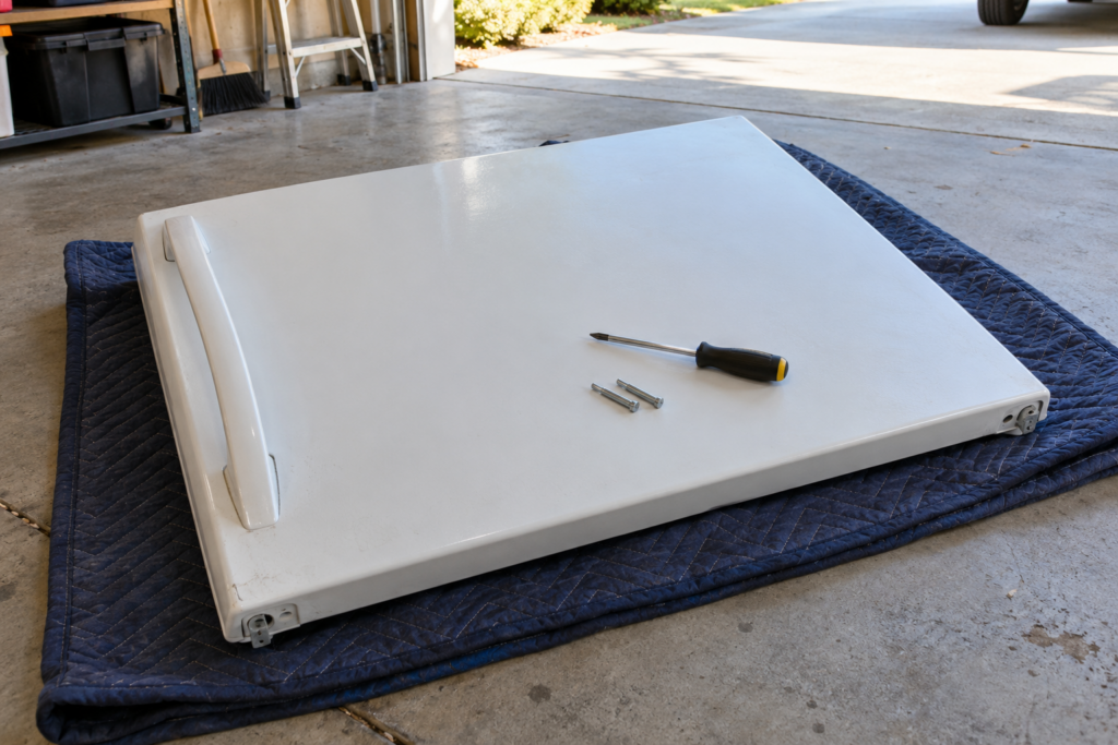 A removed refrigerator door lying on a folded blanket with a screwdriver and hinge pins on top.