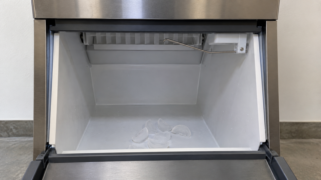 A thin metal arm raised at the top of the open storage bin of an undercounter ice machine with only a few crescent cubes below.